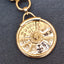 A gold-toned circular zodiac wheel pendant or keychain charm with engraved astrological symbols on a dark textured fabric background.