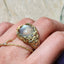 Gold rose ring with a large gemstone held between fingers against a textured background