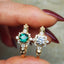 Gold rings with green gemstone and diamond accents held between fingers.