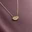 A gold chain necklace with a small crescent-shaped pendant featuring an eye detail, displayed on a textured, dark plum purple fabric background.
