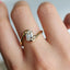 A close-up of a hand wearing a gold ring with a rectangular clear gemstone, set against a soft, blurred light gray background.