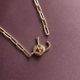 A gold paperclip chain necklace with a small elephant-shaped pendant featuring a red heart and white crystal details, displayed on a textured dark purple background.