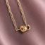 A gold chain necklace with an oval link chain and a padlock-shaped pendant featuring a small red heart in the center, displayed on a textured mauve fabric background.