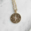 A gold chain necklace with a round pendant featuring a starburst design and small clear stones, displayed on a light marble surface.
